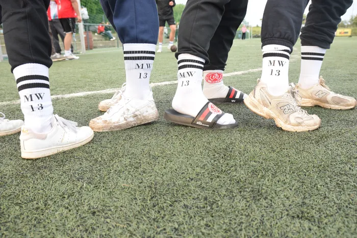 Fußballspieler mit weißen Socken und den Initialen 'MW 13' stehen auf einem grünen Kunstrasenplatz. Einige tragen Sportschuhe, einer hat Badeschlappen an.