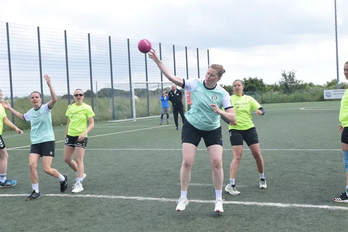 Eine Spielerin wirft einen Ball während eines Handballspiels auf einem grünen Kunstrasenplatz. Im Hintergrund sind weitere Spieler und ein blauer Zaun zu sehen.