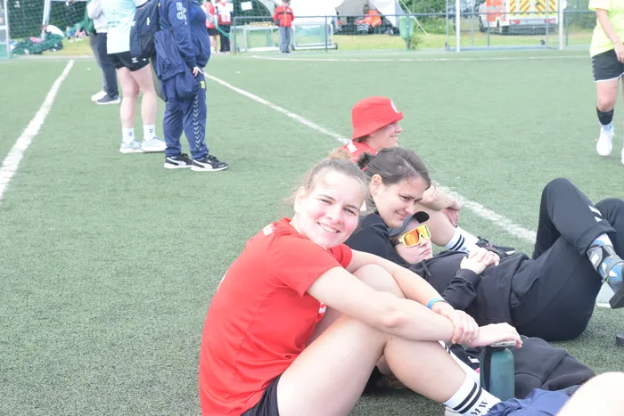 Drei junge Frauen sitzen auf einem grünen Sportplatz, die vorderste lächelt in die Kamera. Im Hintergrund sind weitere Sportler und Zuschauer zu sehen.