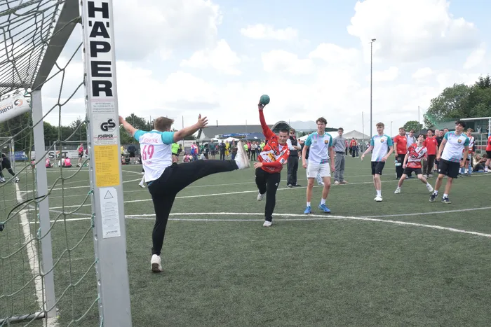Ein Handballspieler schießt den Ball in einem Spiel auf einem grünen Rasenplatz. Im Hintergrund stehen weitere Spieler und Zuschauer.