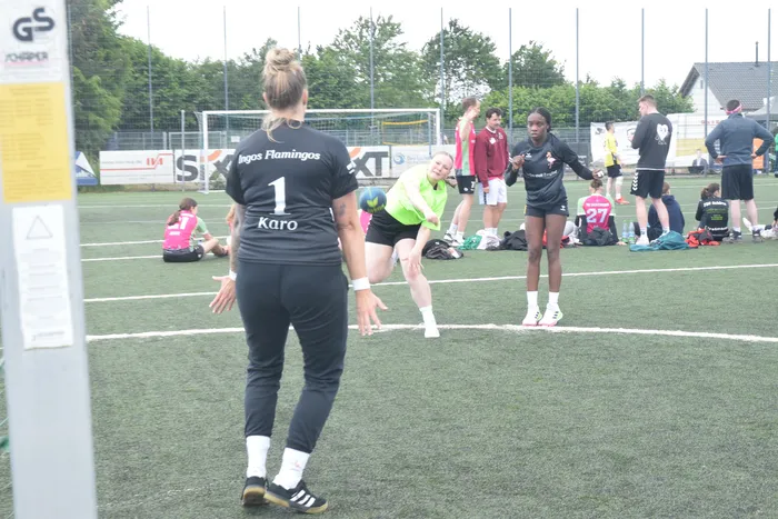 Eine Frau mit kurzen blonden Haaren steht mit dem Rücken zum Betrachter auf einem grünen Rasenplatz. Im Hintergrund stehen weitere Personen, vermutlich Spielerinnen und Trainer, sowie ein Fußballtor.