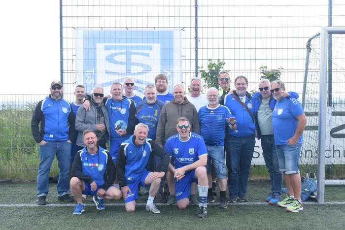 Eine Gruppe von etwa 20 Männern in Sportkleidung, hauptsächlich in Blau, posiert lächelnd auf einem Rasenplatz vor einem blauen Zaun. Einige tragen Trikots mit Vereinslogos und Sponsorenaufdrucken.