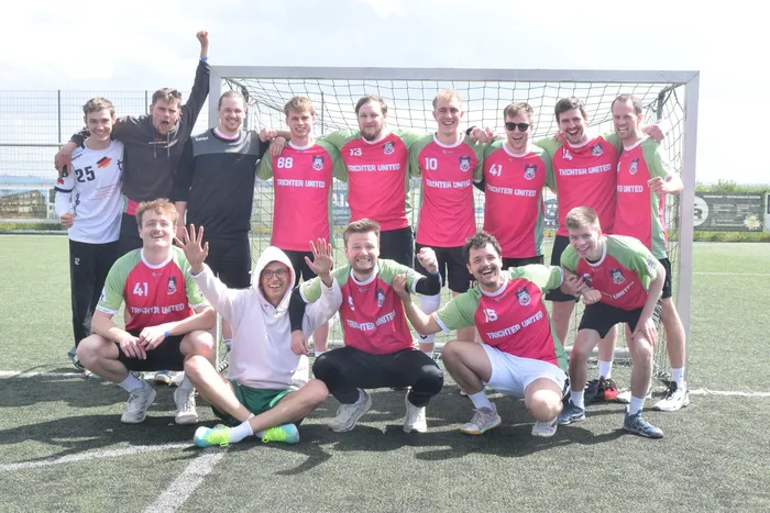 Eine Fußballmannschaft in roten und grünen Trikots mit der Aufschrift „Tüchter United“ posiert vor einem Fußballtor auf einem Kunstrasenplatz. Einige Spieler lächeln und winken in die Kamera.