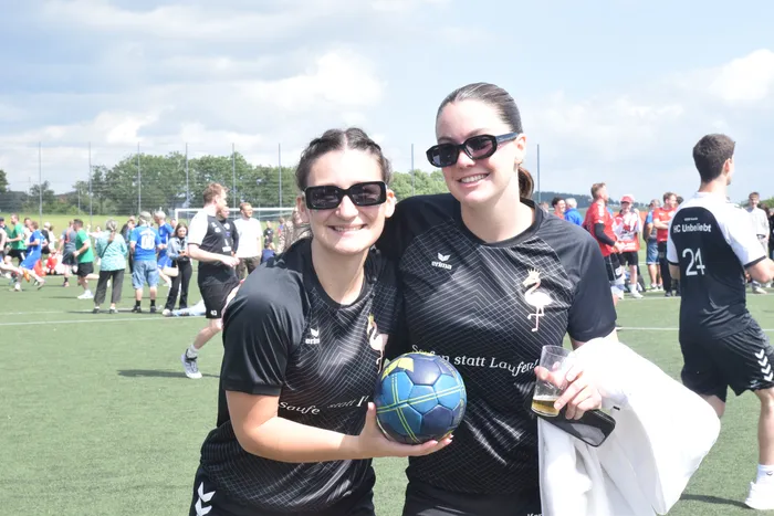 Zwei lächelnde Frauen mit Sonnenbrillen halten einen bunten Handball auf einem grünen Rasenplatz. Im Hintergrund sind weitere Menschen und ein Fußballfeld zu sehen.
