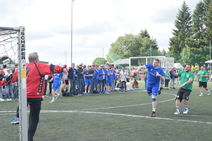 Ein Fußballtorwart in schwarzer und roter Sportkleidung gestikuliert auf einen jubelnden Fußballspieler in blauem Trikot und weißen Shorts, umgeben von weiteren Spielern und Zuschauern auf einem Kunstrasenplatz. Im Hintergrund sind Bäume und Gebäude zu sehen.