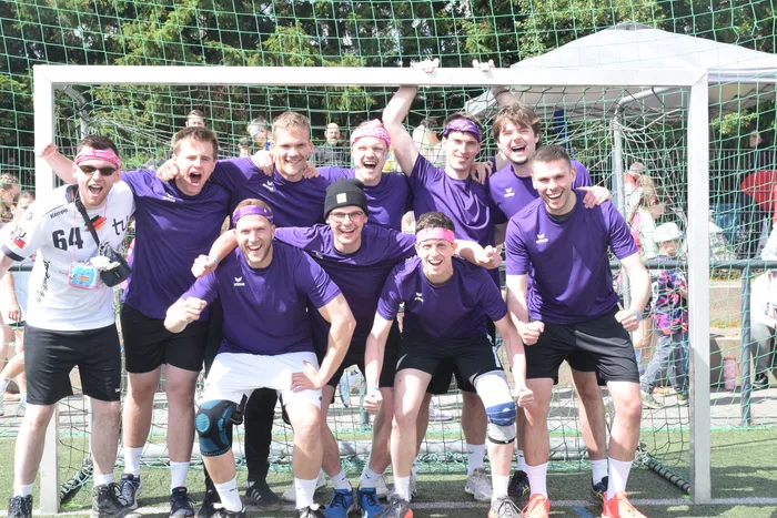 Eine Gruppe junger Männer in violetten Trikots und weißen Shorts jubelt vor einem Handballtor. Einige tragen Stirnbänder und zeigen ihre Freude.