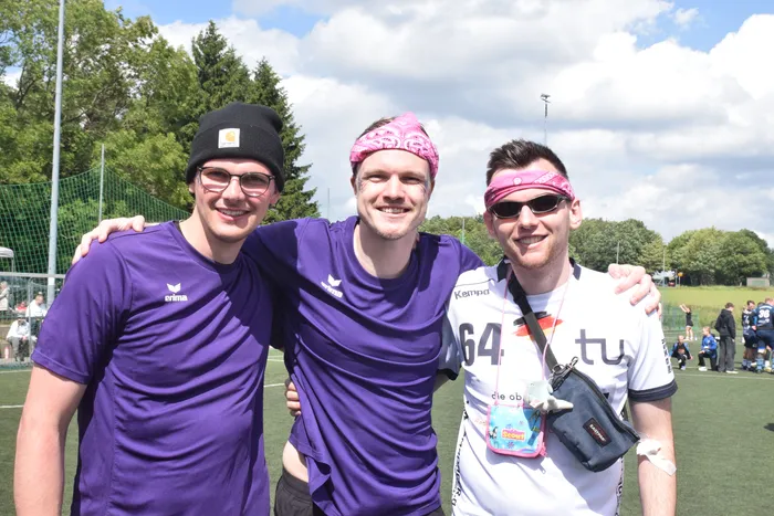 Drei junge Männer mit sportlicher Kleidung stehen eng zusammen und lächeln in die Kamera. Im Hintergrund sind weitere Menschen und ein Sportplatz zu sehen.