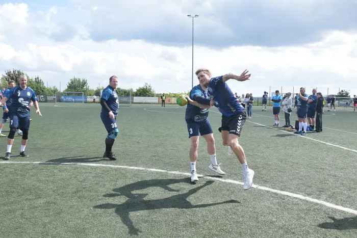 Ein Handballspieler in blauem Trikot springt mit dem Ball in der Hand auf einem grünen Kunstrasenplatz. Im Hintergrund stehen weitere Spieler und Zuschauer.