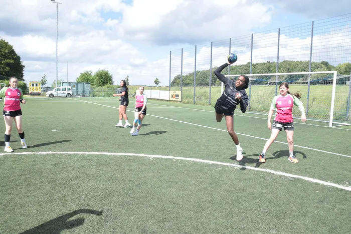 Eine Spielerin wirft im Handballspiel den Ball, während andere Spielerinnen auf dem Feld stehen. Im Hintergrund ist ein Zaun und Bäume zu sehen.