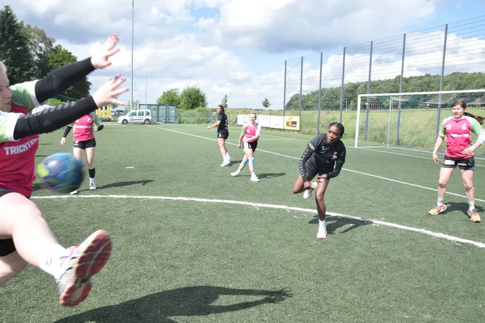 Eine Spielerin in rotem Trikot wirft einen Handball, während andere Spielerinnen auf dem grünen Kunstrasenplatz zusehen. Im Hintergrund ein Handballtor und ein grauer Zaun.