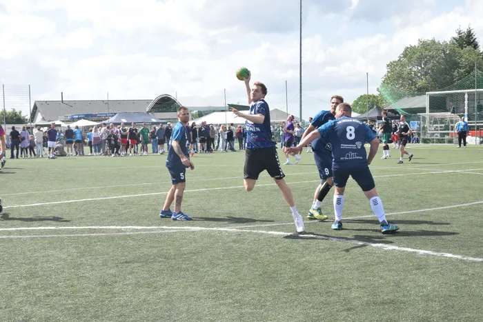 Ein Handballspieler wirft den Ball während eines Spiels auf dem grünen Kunstrasen. Im Hintergrund stehen Zuschauer und sind weitere Spieler zu sehen.