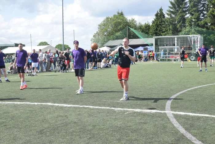 Ein Spieler in roter Hose und weißem Trikot wirft einen Handball auf einem grünen Rasenplatz. Im Hintergrund stehen weitere Spieler und Zuschauer.