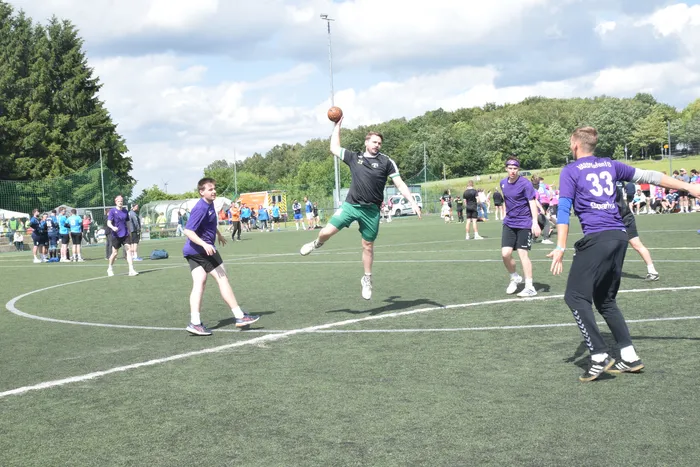 Ein Spieler in schwarzer und grüner Sportkleidung wirft einen Ball während eines Handballspiels auf einem grünen Kunstrasenplatz. Im Hintergrund stehen weitere Spieler und Zuschauer.