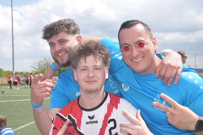 Drei lächelnde Männer in Sportkleidung posieren eng umschlungen für ein Foto. Der Mann in der Mitte trägt ein rotes und weißes Trikot mit der Nummer 2, die beiden anderen blaue Trikots mit dem Aufdruck 