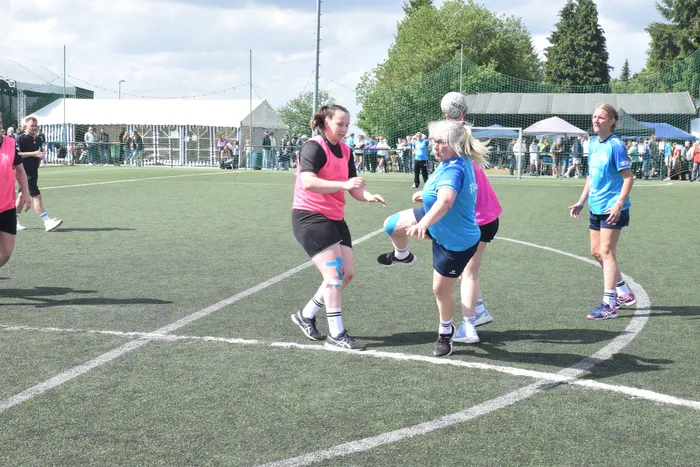 Zwei Frauen in Sportkleidung spielen Handball auf einem grünen Kunstrasenplatz. Im Hintergrund sind weitere Spieler und Zuschauer zu sehen.