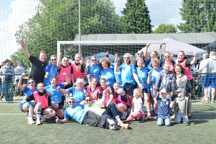 Eine Gruppe von Frauen und Mädchen in Sportkleidung posiert jubelnd vor einem Fußballtor. Einige knien oder hocken, andere stehen und heben die Fäuste in die Luft. Im Hintergrund sind Zuschauer und Zelte zu sehen.