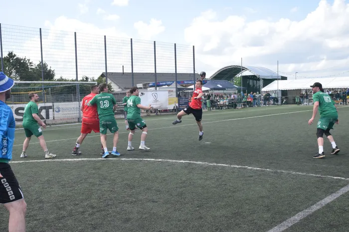 Handballspieler in grünen und roten Trikots kämpfen um den Ball auf einem Kunstrasenplatz. Im Hintergrund sind Zuschauer und ein Stadion zu sehen.