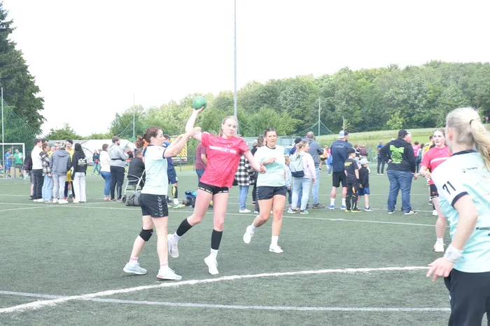 Eine Spielerin im roten Trikot wirft einen Handball während eines Spiels auf einem grünen Kunstrasenplatz. Im Hintergrund sind weitere Spieler und Zuschauer zu sehen.