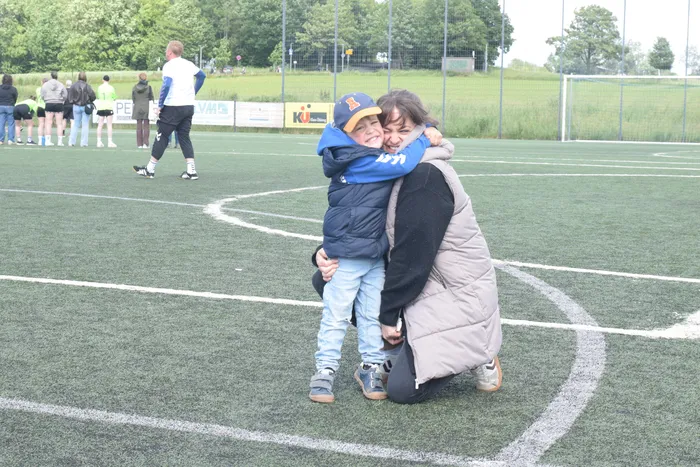 Eine Frau umarmt einen Jungen auf einem grünen Kunstrasenplatz, während im Hintergrund andere Menschen stehen und sich bewegen.