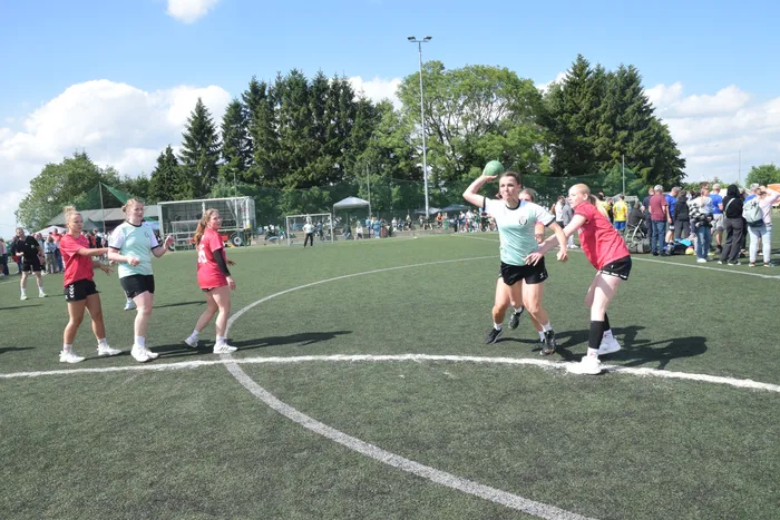 Eine Spielerin wirft einen Handball auf einem grünen Kunstrasenplatz, während andere Spielerinnen in der Nähe stehen und zum Ball laufen. Im Hintergrund sind Zuschauer und Bäume zu sehen.