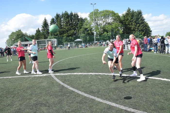 Frauen spielen Handball auf einem grünen Kunstrasenplatz, während Zuschauer im Hintergrund zusehen. Einige Spielerinnen laufen und werfen den Ball.