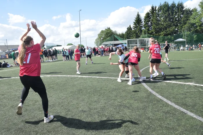Eine Spielerin in einem roten Trikot wirft einen Ball während eines Handballspiels auf einem grünen Kunstrasenplatz. Im Hintergrund stehen weitere Spielerinnen und Zuschauer.
