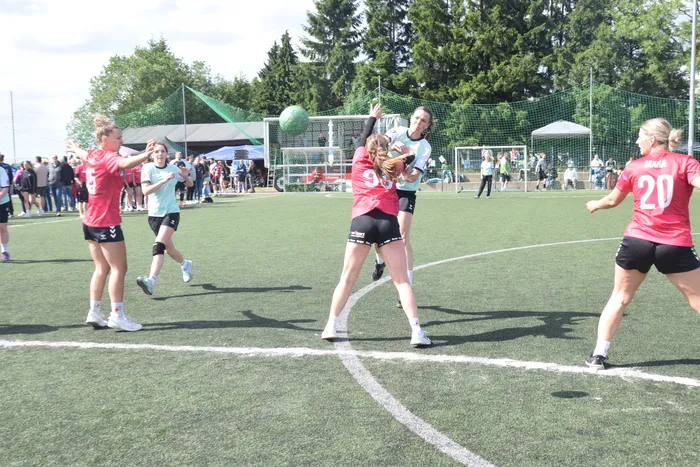 Handballspielerinnen kämpfen um den Ball während eines Spiels auf einem grünen Kunstrasenplatz. Im Hintergrund stehen Zuschauer und ein Handballtor ist zu sehen.