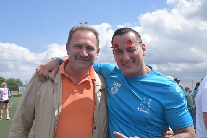 Zwei Männer lächeln in die Kamera, der Mann rechts trägt ein hellblaues Handballtrikot mit dem Aufdruck 