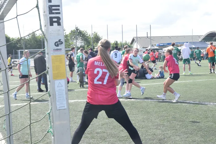 Eine Spielerin mit der Rückennummer 27 im roten Trikot steht im Vordergrund eines Handballfeldes, während im Hintergrund weitere Spieler und Zuschauer zu sehen sind. Im Hintergrund sind auch Werbebanner und ein Stadion zu erkennen.