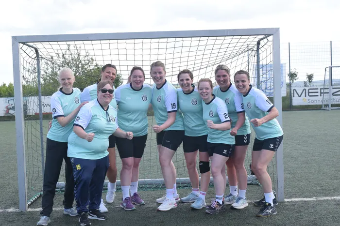 Eine Gruppe von acht Frauen in hellgrünen Handballtrikots und Trainingsanzügen steht lächelnd vor einem Fußballtor auf einem Kunstrasenplatz. Im Hintergrund ist ein Werbebanner zu sehen.