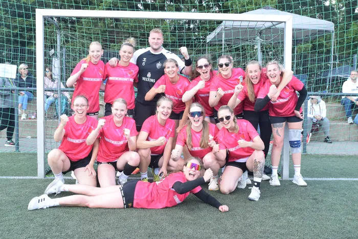Eine Gruppe von jungen Frauen in roten Trikots und schwarzen Shorts posiert jubelnd vor einem Fußballtor. Einige zeigen Daumen hoch oder Faustzeichen, eine Person liegt lachend im Vordergrund.