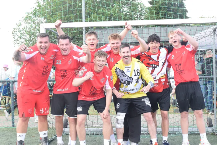 Eine Gruppe von jungen Männern in Sportkleidung posiert jubelnd vor einem Fußballtor. Einige tragen rote Trikots mit der Nummer 23 und 1, ein anderer ein gelbes Trikot mit der Nummer 18.