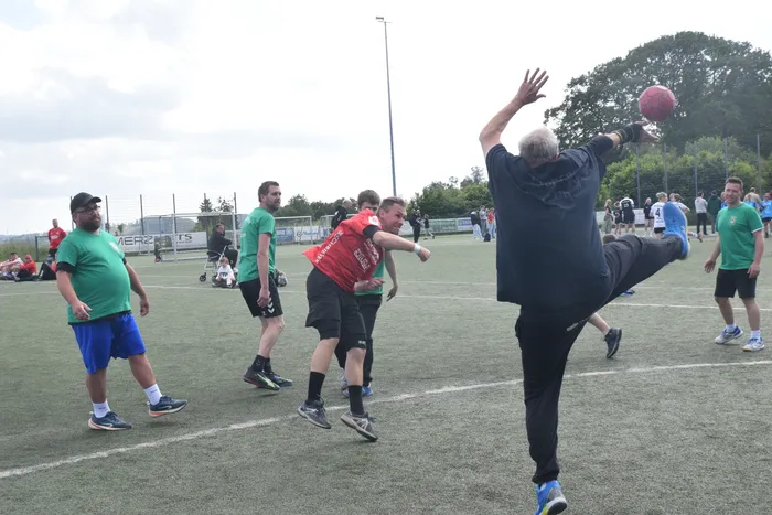Ein Spieler in schwarzer Hose und Trikot springt beim Handballspiel hoch, um den Ball zu fangen. Im Hintergrund sind weitere Spieler und Zuschauer auf dem grünen Spielfeld zu sehen.