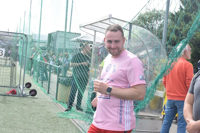 Ein Mann mit kurzem, braunem Haar hält ein Mikrofon in der Hand und steht vor einem Tor auf einem Sportplatz. Er trägt ein hellrosa Trikot mit Werbeaufdruck und rote Shorts. Im Hintergrund ist ein grauer Hügel zu sehen.
