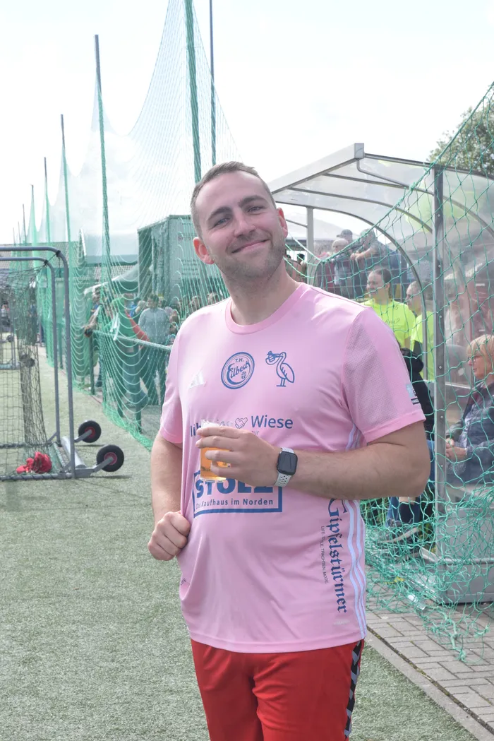 Ein Mann mit braunen Haaren und Bart lächelt in die Kamera. Er trägt ein hellrosa T-Shirt mit dem Logo von „Wiese“ und „Stolzenhagen“ sowie Trainingshose und Sneaker. Im Hintergrund sind Netze und weitere Personen zu sehen.