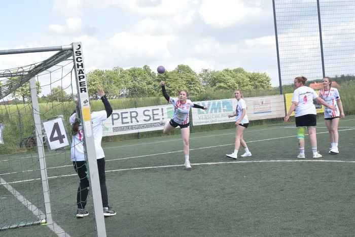 Eine Torhüterin springt nach einem Ball, der von einer Spielerin in rotem Trikot geworfen wurde. Im Hintergrund stehen weitere Spielerinnen auf dem Spielfeld. Im Hintergrund sind Werbebanner zu sehen.