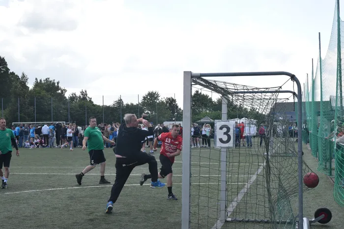 Handballspieler in Aktion während eines Turniers auf einem grünen Feld. Ein Spieler in rotem Trikot versucht, den Ball ins Tor zu werfen, während ein Gegenspieler in Schwarz ihn blockt. Im Hintergrund stehen Zuschauer und weitere Spieler.