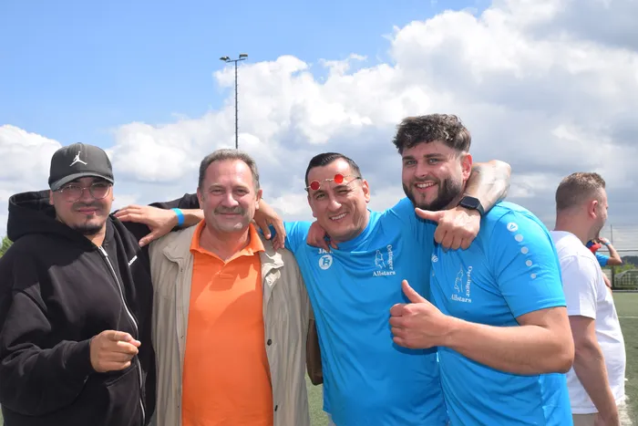 Vier Männer stehen lächelnd eng zusammen vor einer sportlichen Kulisse. Zwei tragen blaue Sporttrikots mit dem Aufdruck 