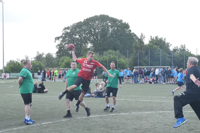 Ein Spieler in rotem Trikot wirft einen Handball während eines Spiels auf einem grünen Feld. Im Hintergrund stehen und sitzen Zuschauer, während weitere Spieler in grünen Trikots agieren.