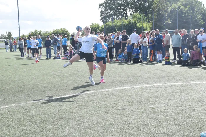 Eine Spielerin wirft den Ball während eines Handballspiels auf einem grünen Kunstrasenplatz. Im Hintergrund stehen Zuschauer und weitere Spielerinnen und Spieler.