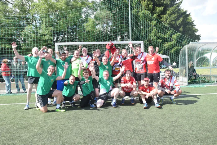 Eine Gruppe von etwa 20 Personen, die meisten in Sportkleidung, posiert jubelnd vor einem Fußballtor auf einem Kunstrasenplatz. Die Spieler tragen Trikots in Grün und Rot.