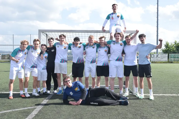 Eine Gruppe junger Männer in Handballtrikots steht vor einem Tor und jubelt. Ein Teammitglied liegt lächelnd vor der Gruppe. Im Hintergrund sind Werbebanner zu sehen.