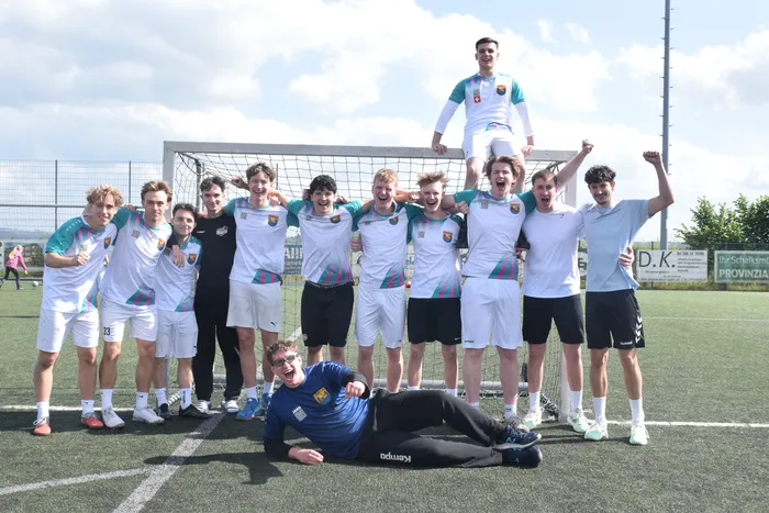 Eine Fußballmannschaft steht vor einem Tor auf einem Kunstrasenplatz. Ein Spieler liegt vor der Gruppe, während ein anderer auf dem Tornetz steht und die Arme hebt.