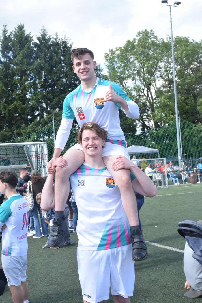 Zwei junge Männer in Handballtrikots mit Schweizer Flagge lächeln in die Kamera. Der vordere trägt den anderen auf seinen Schultern, im Hintergrund sind weitere Spieler und Zuschauer zu sehen.