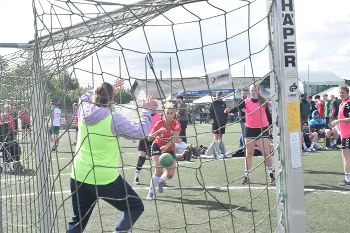 Eine Spielerin in Weste versucht, den Ball vor der Torwartin in rotem Trikot zu blocken, während weitere Spielerinnen im Hintergrund stehen. Im Hintergrund sind Zuschauer und Werbebanner zu sehen.