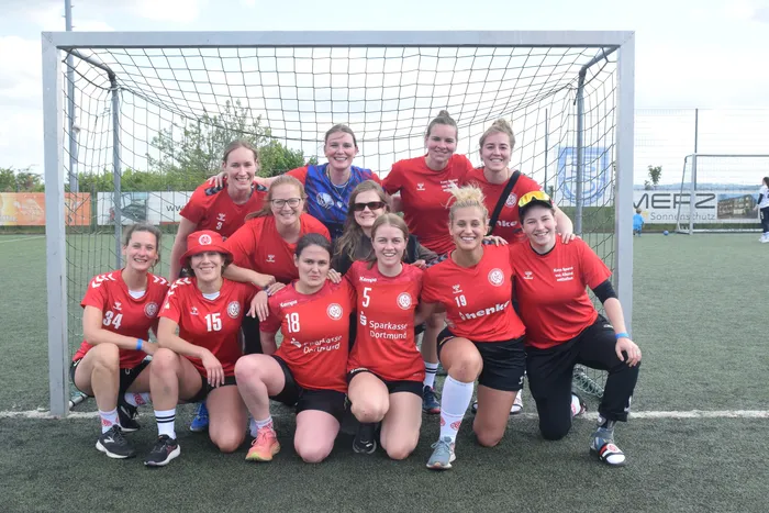 Eine Gruppe von elf Frauen in roten Trainingsanzügen und einigen mit Basecaps posiert vor einem Fußballtor. Die Sportlerinnen lächeln in die Kamera, einige tragen Sonnenbrillen. Im Hintergrund ist ein Fußballfeld und ein Werbebanner zu sehen.