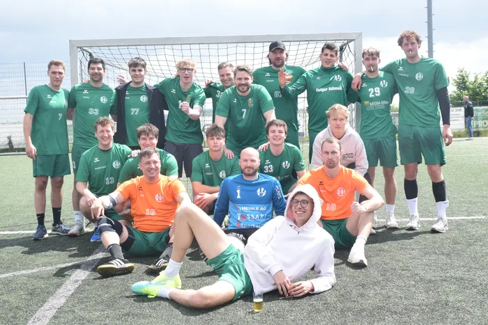 Eine Gruppe von jungen Männern in Sportkleidung posiert vor einem Fußballtor auf einem Kunstrasenplatz. Die meisten tragen grüne Trikots mit der Nummer und dem Logo eines Sponsors, einige orangefarbene Trikots. Einige halten Bierflaschen oder -gläser in der Hand.