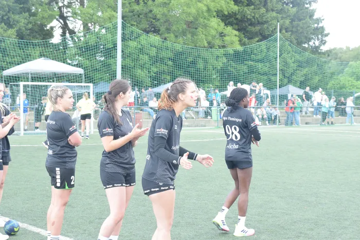 Fünf Frauen in schwarzen Handballtrikots stehen auf einem grünen Kunstrasenplatz. Zwei Spielerinnen geben sich die Hände, während die anderen zusehen. Im Hintergrund sind weitere Menschen und ein Zaun zu erkennen.