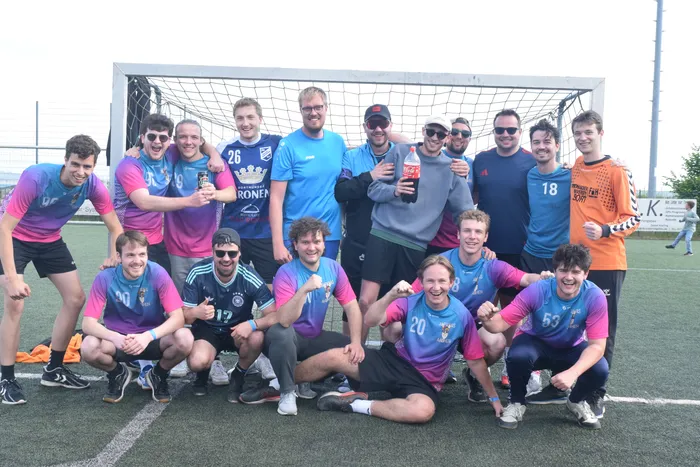 Eine Gruppe junger Erwachsener in Sportkleidung posiert lächelnd vor einem Fußballtor auf einem Kunstrasenplatz. Einige halten Getränke in der Hand.