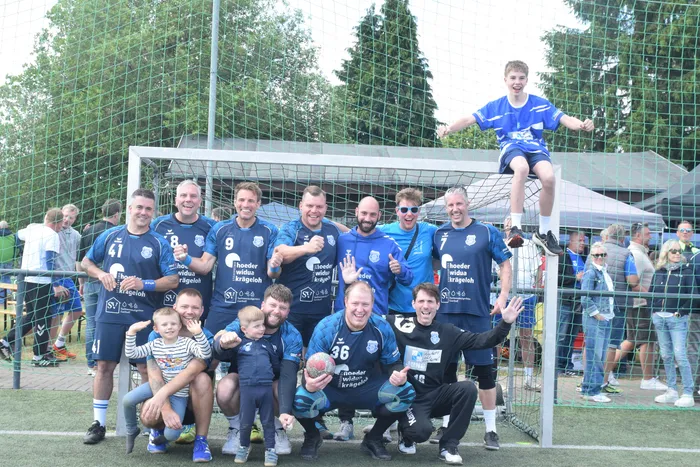 Eine Handballmannschaft in blau-weißen Trikots posiert vor einem Tor auf einem grünen Rasenfeld. Ein Spieler steht auf dem Torpfosten, während die anderen Spieler und Kinder um ihn herum stehen und lächeln. Im Hintergrund sind Zuschauer zu sehen.